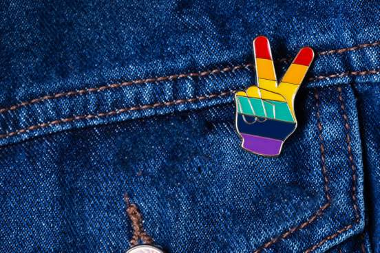 photo of an enamel pin on denim the pin is in the shape of a hand showing a peace sign colored with the rainbow flag 