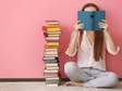 woman sitting on floor holding book in front of her face on pink background