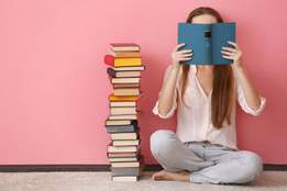 woman sitting on floor holding book in front of her face on pink background